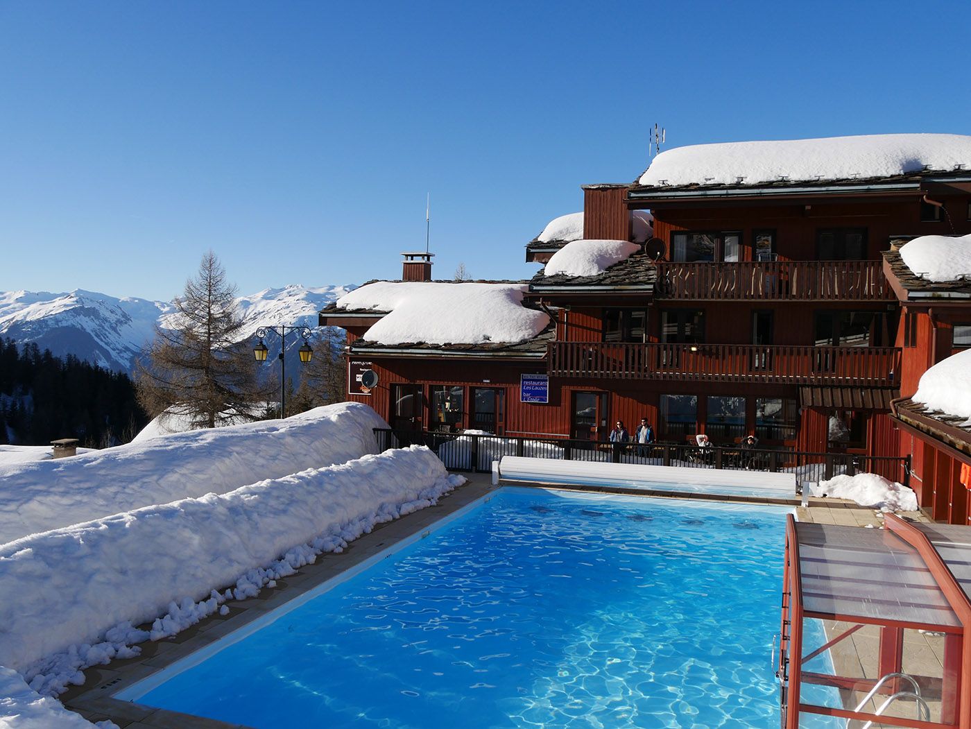 A hotel with a pool surrounded by snow, a mountain view, and a sign indicating a restaurant.