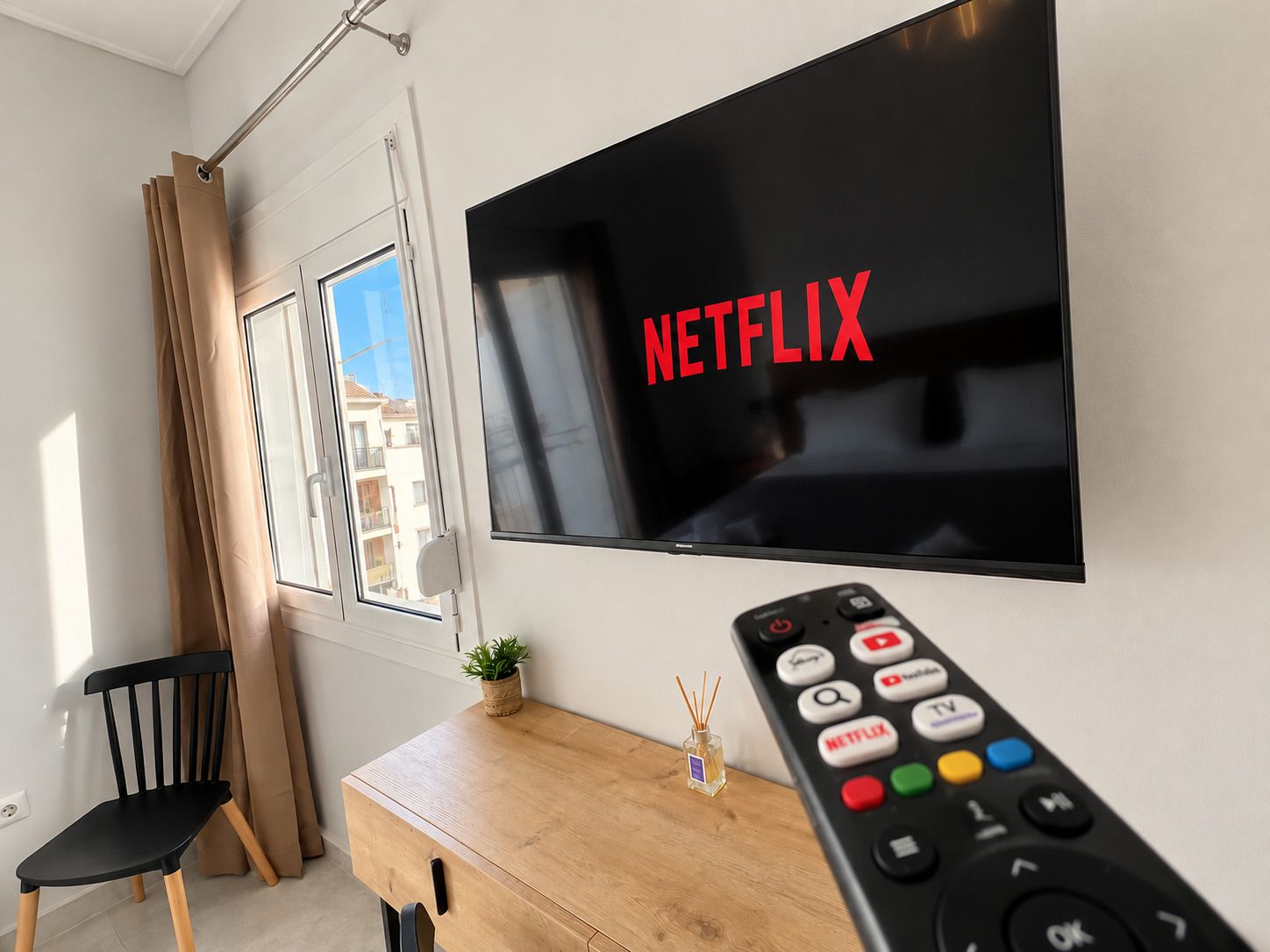 Un salon moderne avec un grand téléviseur à écran plat fixé au mur, affichant le mot « NETFLIX » en lettres rouges, un bureau en bois avec une télécommande, une chaise noire et une fenêtre avec des rideaux.