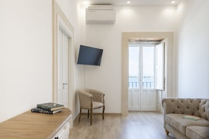 A room with a flat-screen TV mounted on the wall, a wooden table with books, a chair, and a sofa. - Ortigia Prestige | 2 bedrooms and 2 bathrooms, just a short walk from the Duomo (Siracusa)