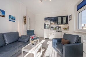 Ein modernes Wohnzimmer mit einem grauen Sofa, einem Couchtisch und einem blauen Sessel. Im Hintergrund ist eine Küche mit weißen Schränken und einem eingebauten Backofen zu sehen.