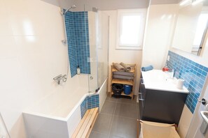 A bathroom with a glass-enclosed shower, a wooden bench, a sink with a mirror, and a window providing natural light. - Large family villa on the beach by Groom* (Larmor-Plage)