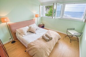 Een slaapkamer met een bed, nachtlampen, een nachtkastje, een raam met uitzicht op zee en een klein tafeltje.