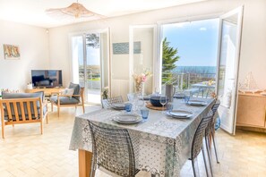 A dining area with a table set for four, a television in the corner, and a view of the sea through large glass doors. - Large family villa on the beach by Groom* (Larmor-Plage)
