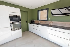 A modern kitchen with white cabinetry, a built-in oven, a coffee maker, and a window with blinds. - Stunning premium family villa, within walking distance of the beach – by Groom* (Guidel)
