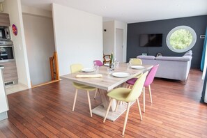 A modern dining area with a wooden table and chairs, a flat-screen TV mounted on the wall, and a window providing natural light. - Modern beachfront house by Groom* (Larmor-Plage)