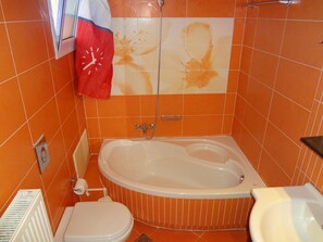 A bathroom with an orange tiled wall, a white bathtub, a white toilet, and a red clock on the wall. - Valena Studios Apartments (Lefkada)
