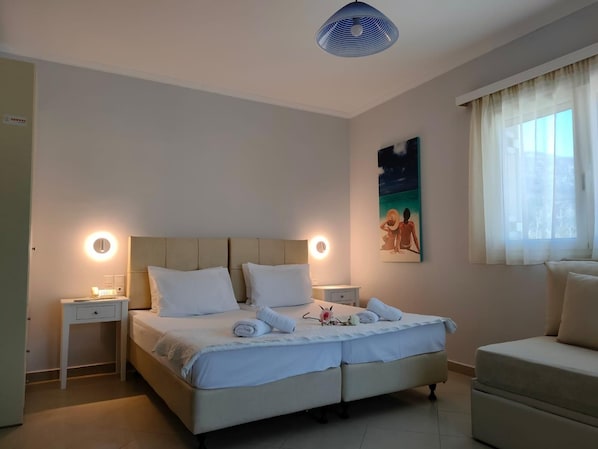 A bedroom with a bed, bedside tables, a nightstand, a chair, a painting, and a window with curtains. - Valena Studios Apartments (Lefkada)