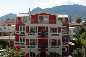 A multi-story building with balconies, a red and white color scheme, and a mountainous backdrop. - Valena Studios Apartments (Lefkada)