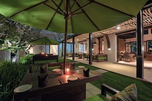 An outdoor dining area with a green umbrella, tables, chairs, and a grassy lawn. - Kemangi Bed and Breakfast (Kuta)