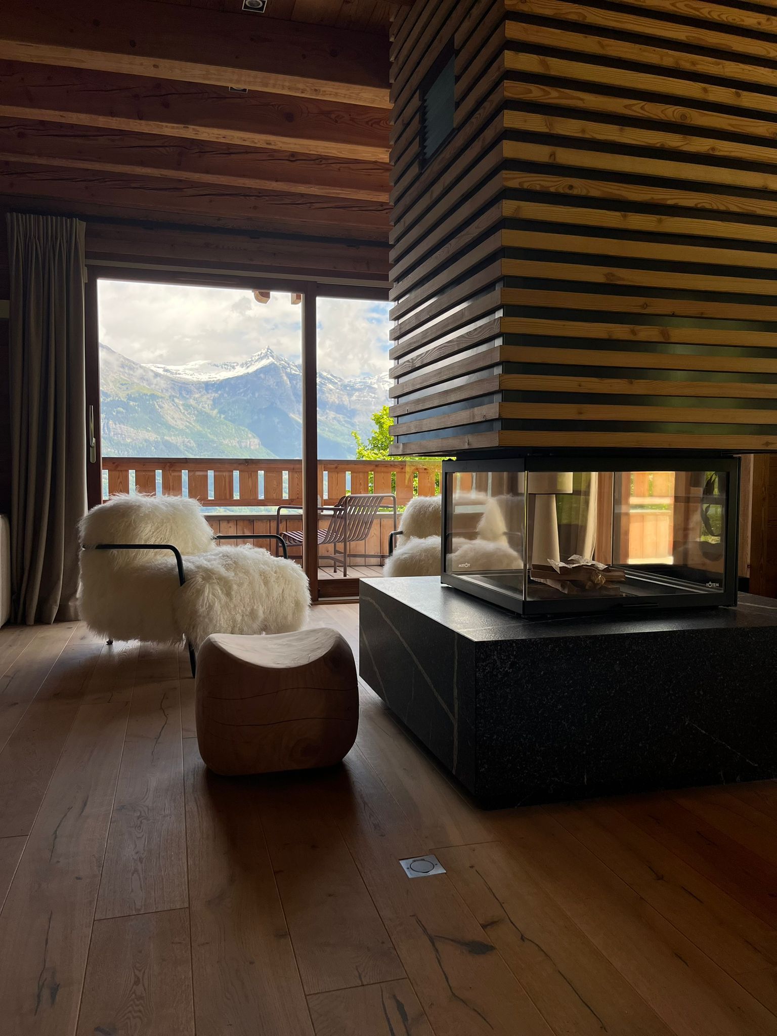 A cozy room with a wooden ceiling, a large window with a mountain view, a fireplace, and comfortable seating.
