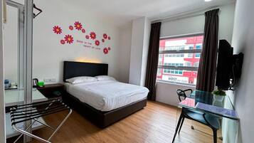 A hotel room with a bed, a desk, a chair, a window with curtains, and a wall with a flower decal.