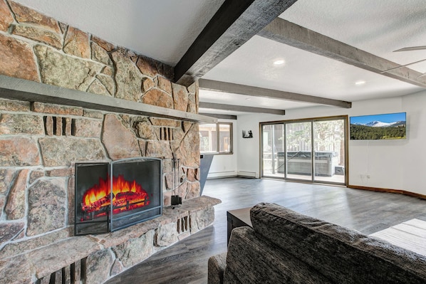 A modern living room with a stone fireplace, a large window, and a flat-screen TV mounted on the wall. - Golf Course Retreat | Hot Tub, Mtn Views, 5BR (Estes Park)
