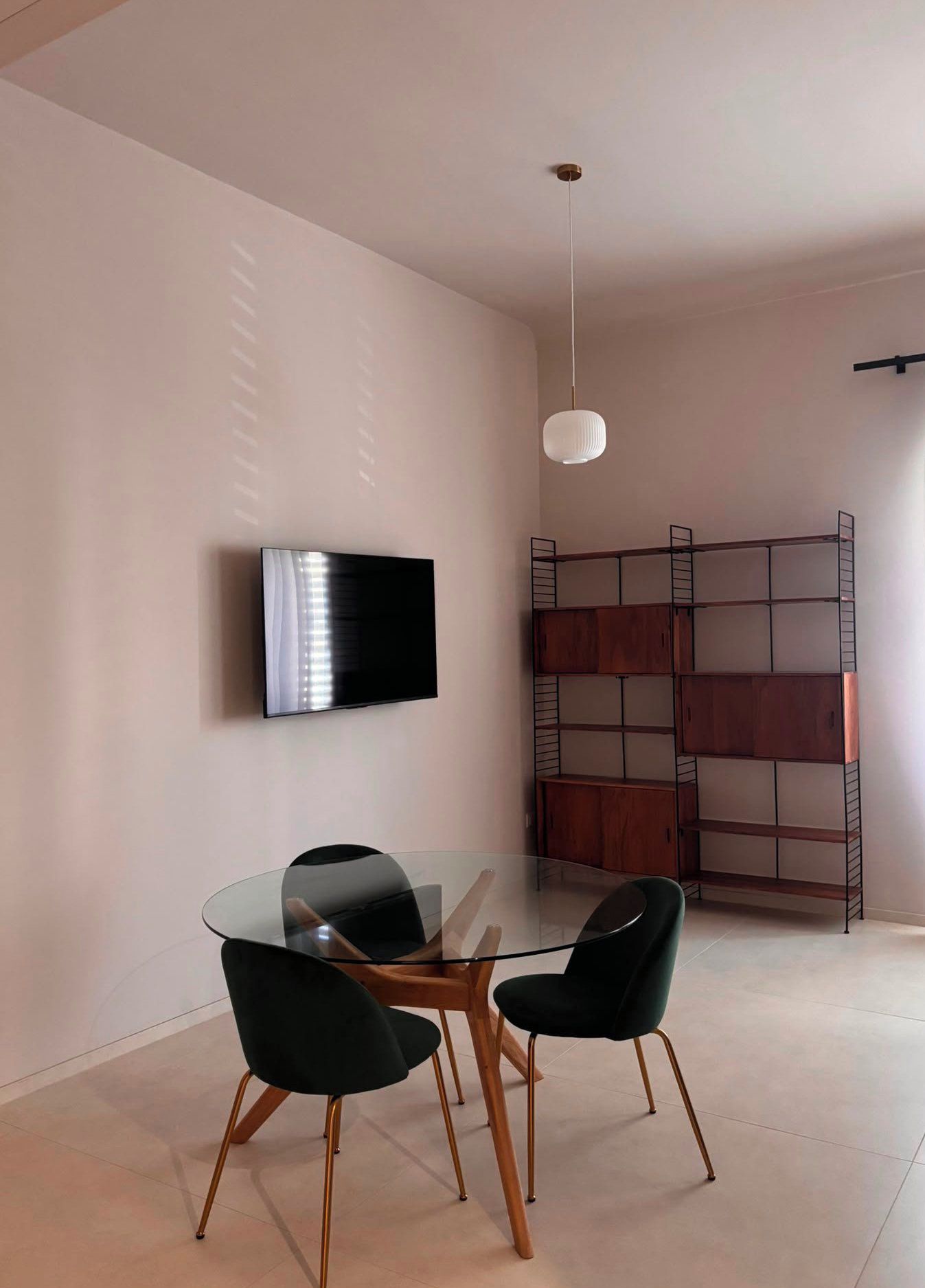 A modern room with a flat-screen TV mounted on the wall, a glass dining table with two chairs, and a metal shelving unit with cabinets.