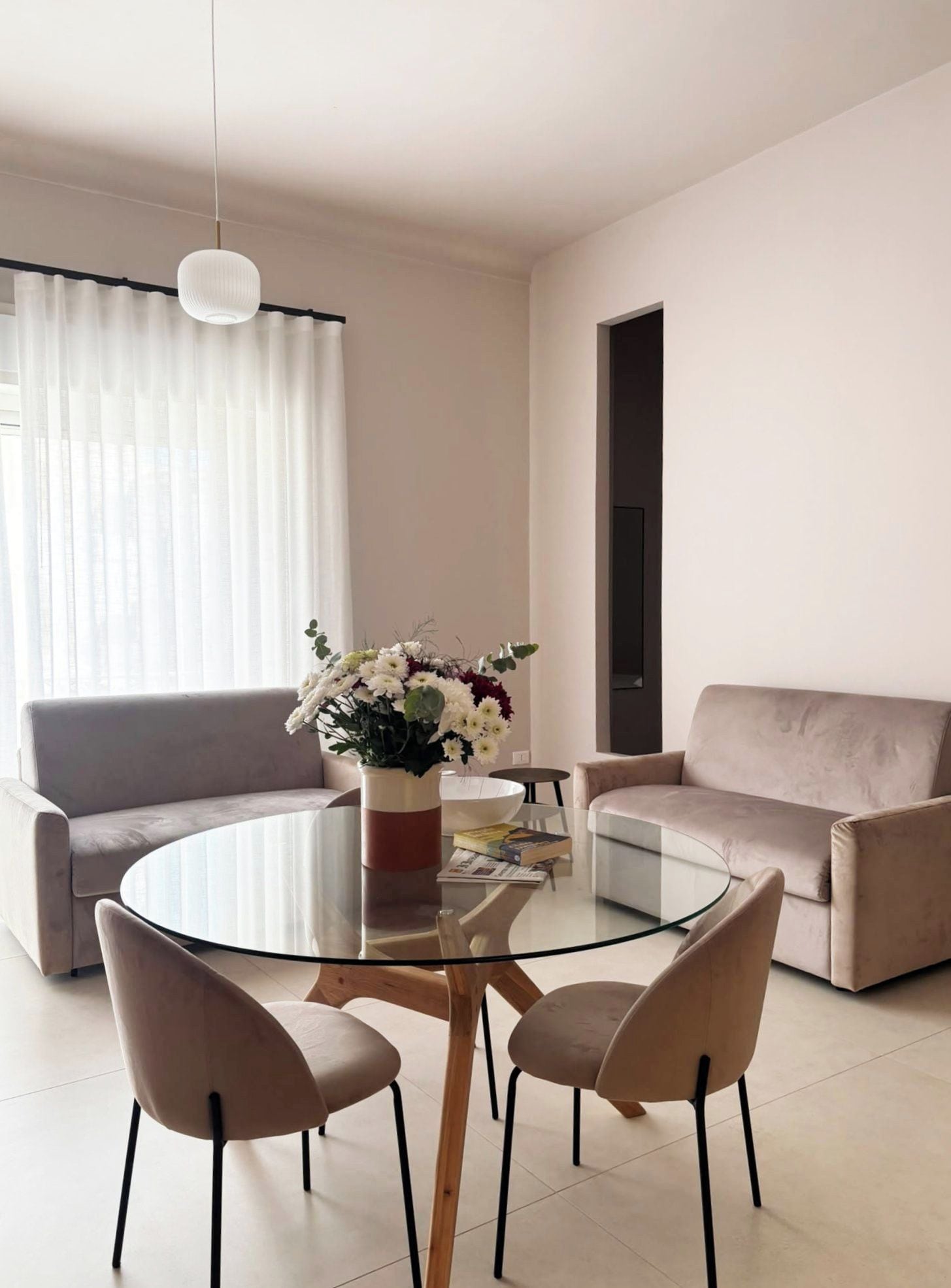 Ein modernes Wohnzimmer mit einem Glastisch, zwei Stühlen, einem Sofa und einer dekorativen Vase mit Blumen.