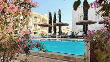 A swimming pool with lounge chairs and umbrellas, surrounded by buildings and flowering plants.