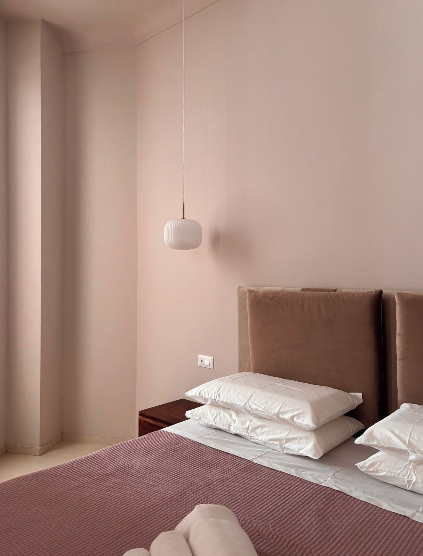 Een slaapkamer met een bed, kussens, een nachtkastje, een hanglamp en een raam met gordijnen.