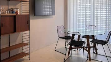 A modern dining area with a glass table and metal chairs, a TV mounted on the wall, and a ceiling light fixture.