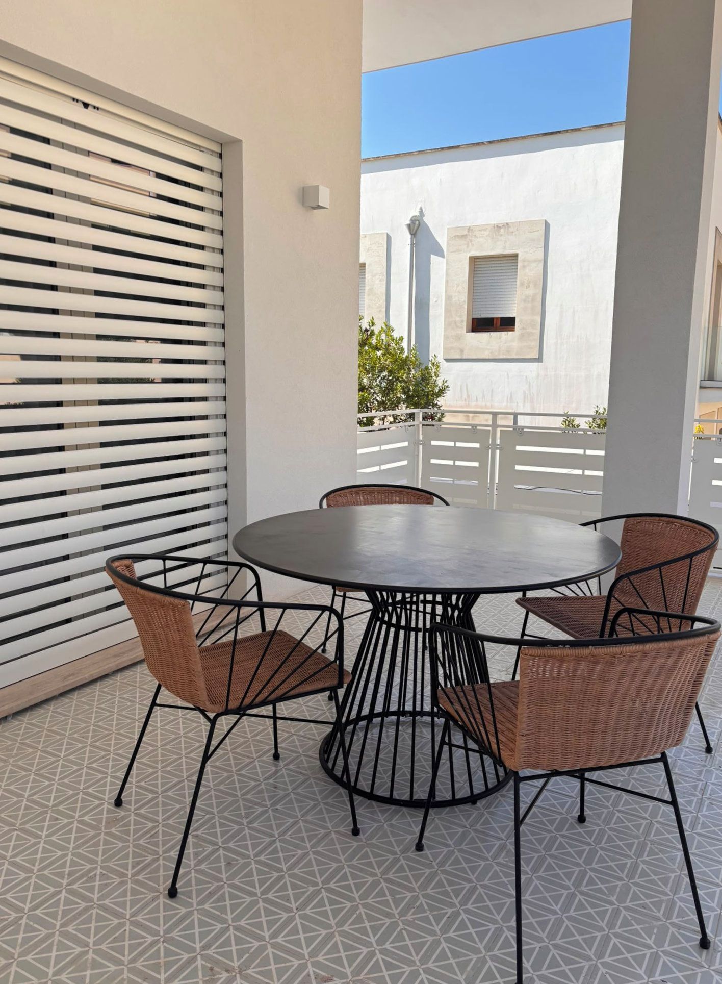 Une table ronde entourée de quatre chaises, un mur blanc avec une fenêtre et un sol orné d’un motif géométrique.