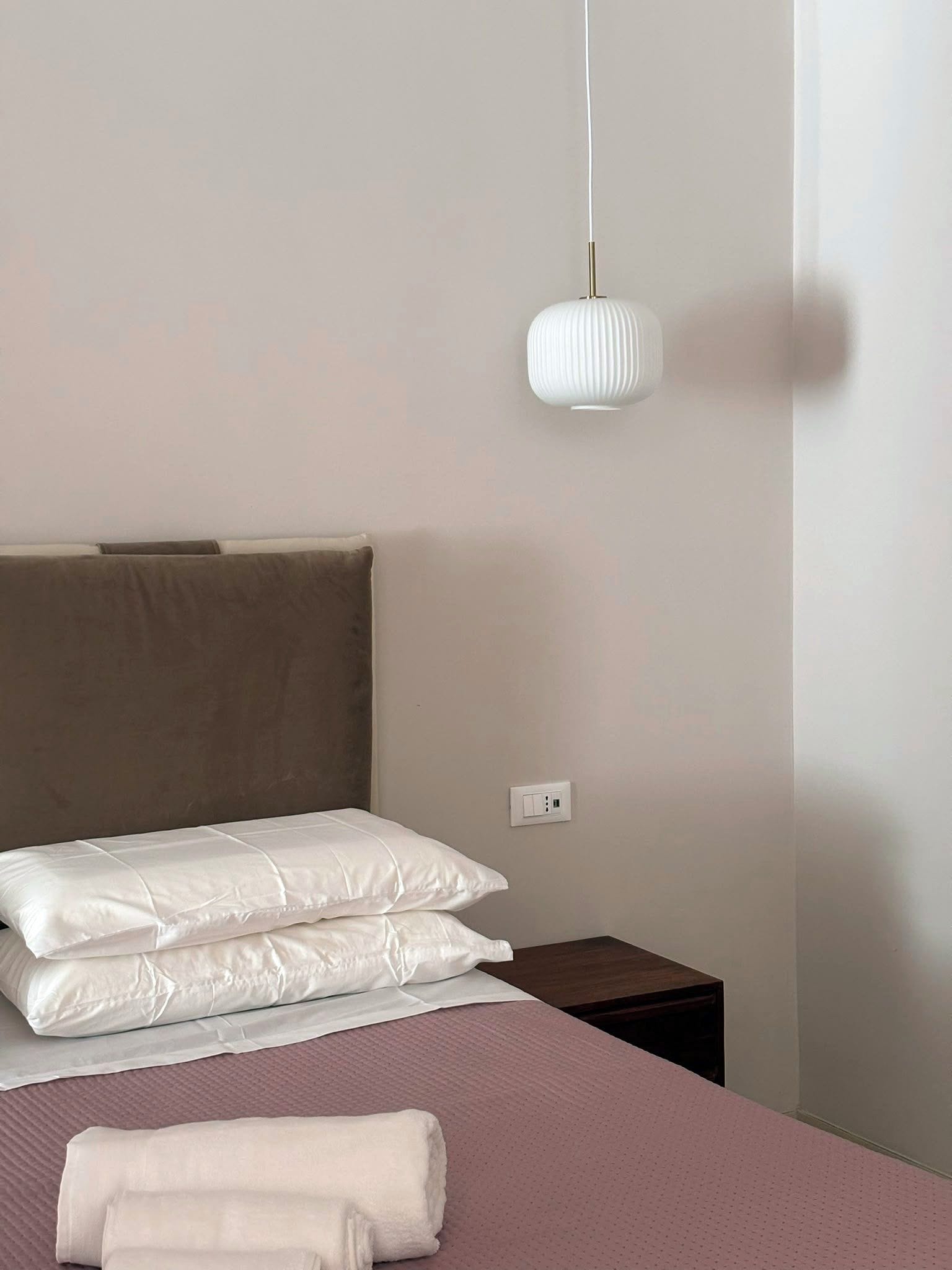 Una cama bien hecha con ropa de cama blanca, un cabecero marrón, una mesita de noche y una lámpara colgante blanca.