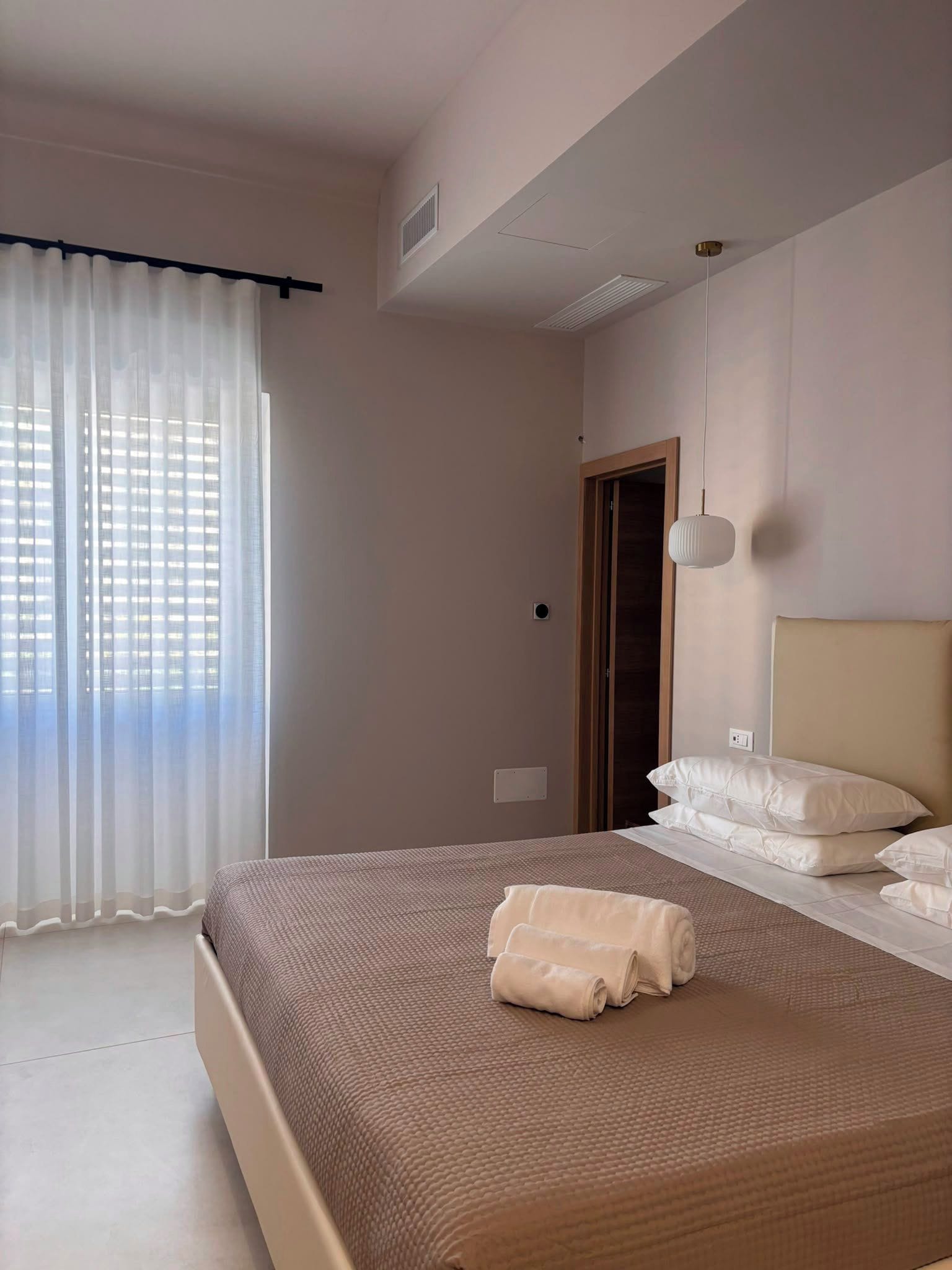 Un dormitorio con una cama grande, una ventana con cortinas translúcidas, una puerta y una lámpara fijada a la pared.