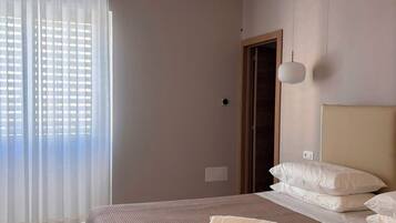 Un dormitorio con una cama grande, una ventana con cortinas translúcidas, una puerta y una lámpara fijada a la pared.