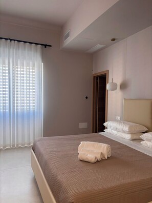 Un dormitorio con una cama grande, una ventana con cortinas translúcidas, una puerta y una lámpara fijada a la pared.