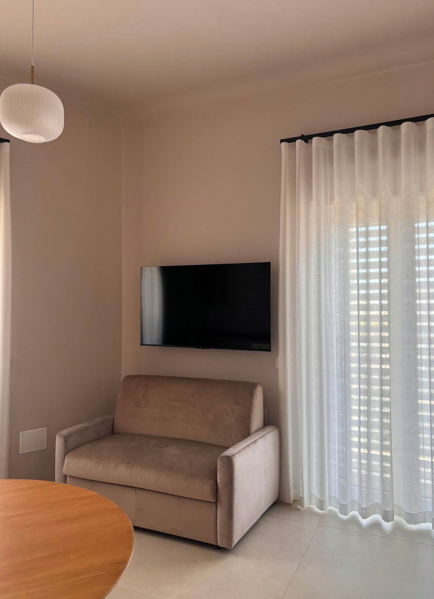 Una sala de estar con un televisor de pantalla plana montado en la pared, un sofá color beige, una mesa redonda de madera y cortinas traslúcidas en la ventana.