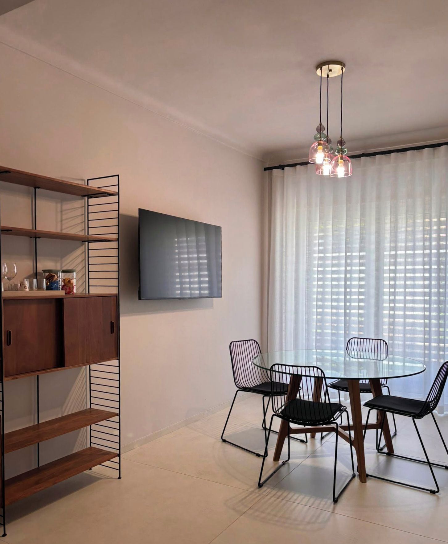 A modern dining area with a glass table and metal chairs, a TV mounted on the wall, and a ceiling light fixture.