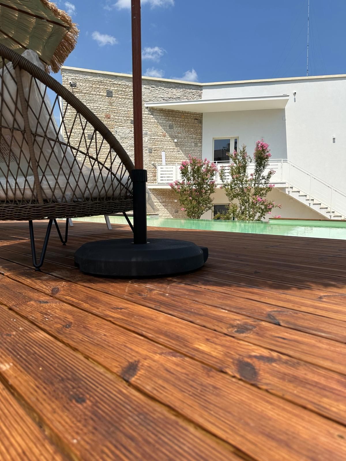 A wooden deck with a chair and a table, a stone wall, a modern building, and a small garden with pink flowers.