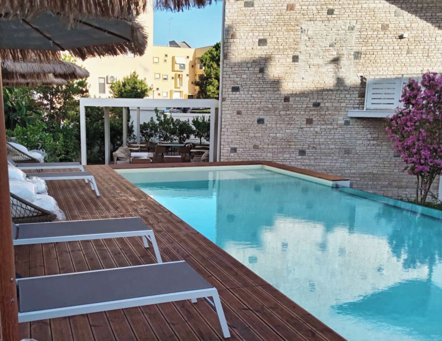 Una piscina con cubierta de madera y sillas de descanso, rodeada por un muro de ladrillo y un edificio con balcones.