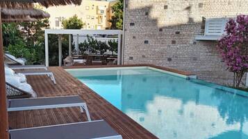 Una piscina con cubierta de madera y sillas de descanso, rodeada por un muro de ladrillo y un edificio con balcones.