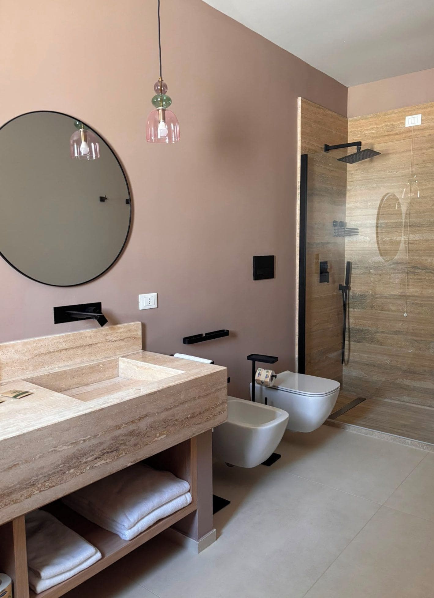 A modern bathroom with a large mirror, a round light fixture, a shower enclosure, and a vanity with a sink and towel storage.
