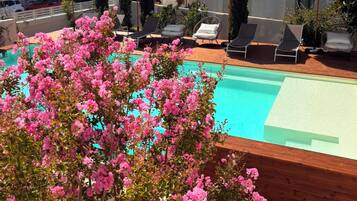 Un espace piscine aménagé sur le toit, avec des chaises longues et des parasols, entouré de plantes à fleurs roses.