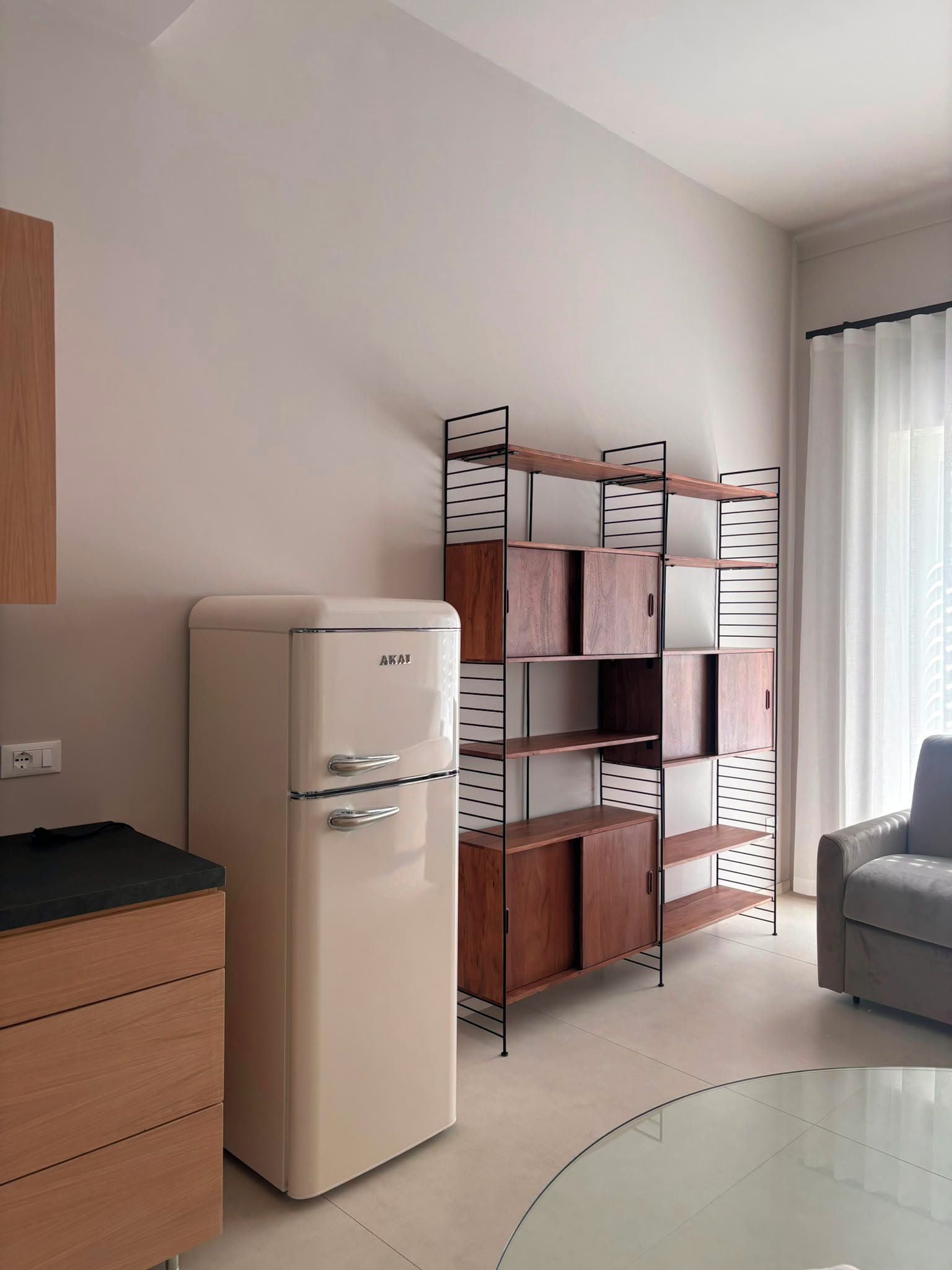 Eine kleine Küchenzeile mit Kühlschrank, einem Holzschrank und einem Metallregal an einer schlichten Wand.