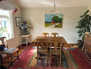 A dining area with a wooden table and chairs, a colorful rug, a painting on the wall, and a potted plant.