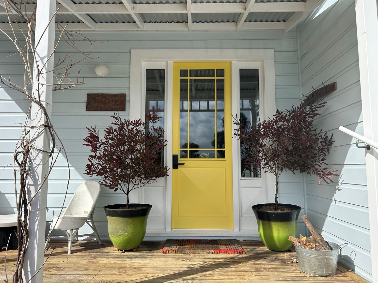 A porch with a yellow door, two potted plants, a white chair, and a metal bucket with logs.
