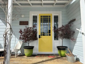 A porch with a yellow door, two potted plants, a white chair, and a metal bucket with logs.