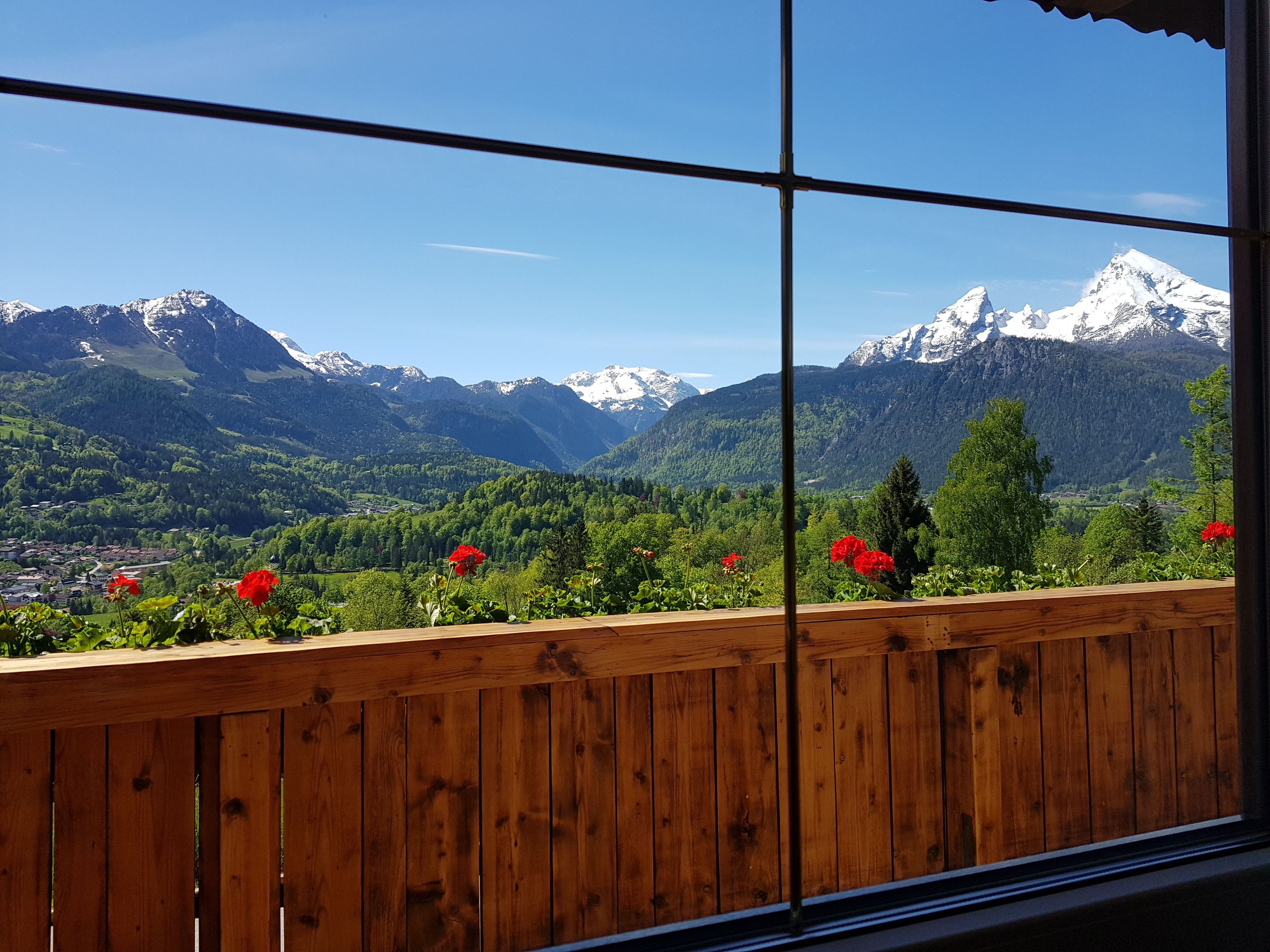 A view through a window with a wooden railing, overlooking a mountainous landscape with snow-capped peaks and lush greenery.