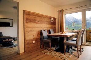 A dining area with a wooden table, chairs, and a view of mountains through a window. - Kälbersteinstüberl Vacation Rental (Bischofswiesen)