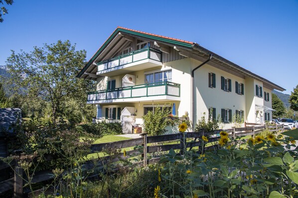 Un bâtiment à deux étages doté d’un balcon, entouré de verdure et d’une clôture en bois.