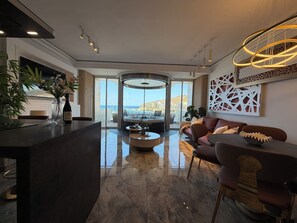 Un soggiorno moderno con un tavolino al centro, un divano e un'area pranzo. Una grande finestra offre vista sul mare e sugli edifici.