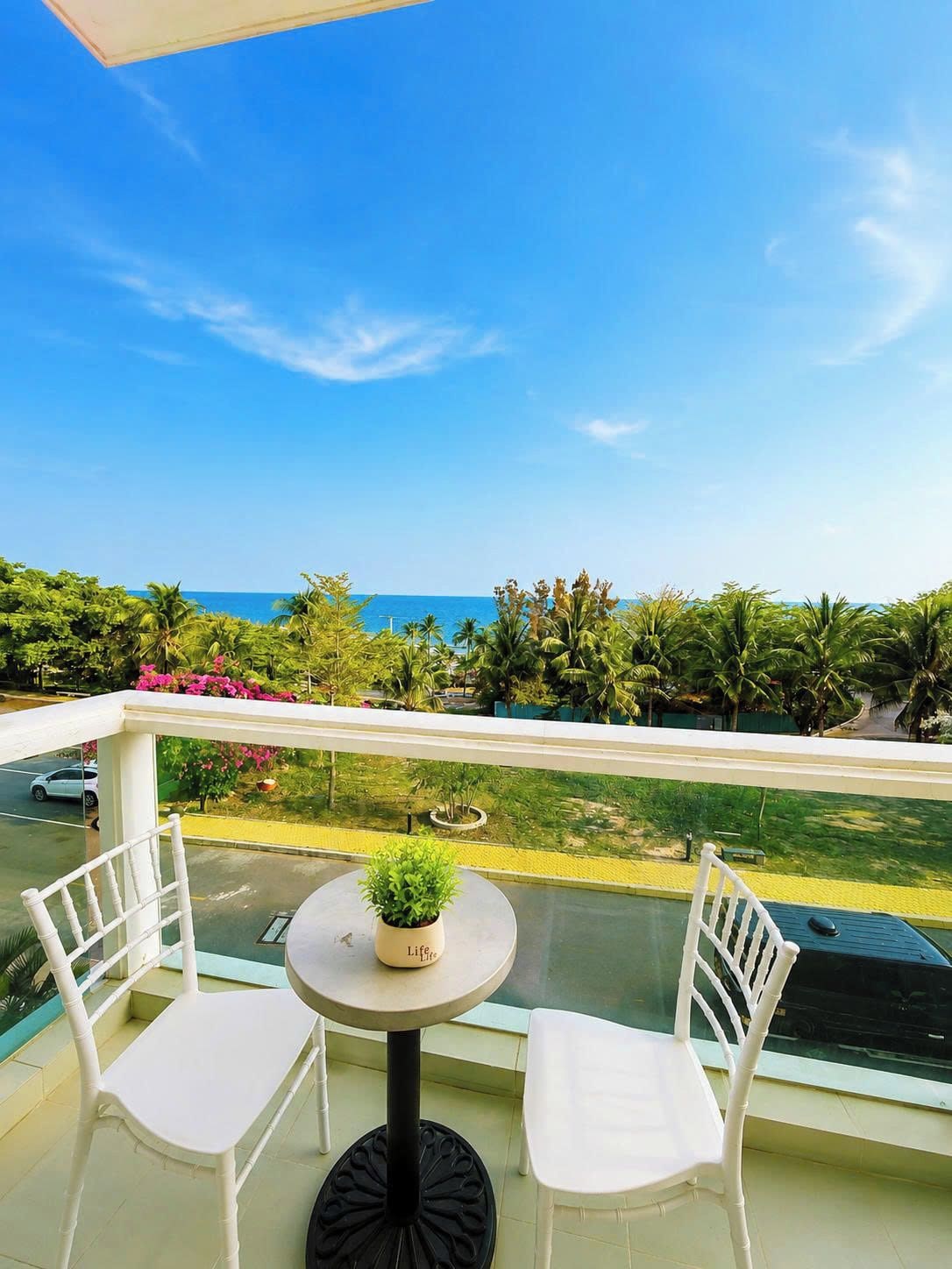 A balcony with two white chairs, a small round table with a potted plant, and a view of the sea and greenery.