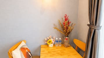 A small dining area with a wooden table and chairs, a wall-mounted light fixture, a vase with dried flowers, and a bottle of water.