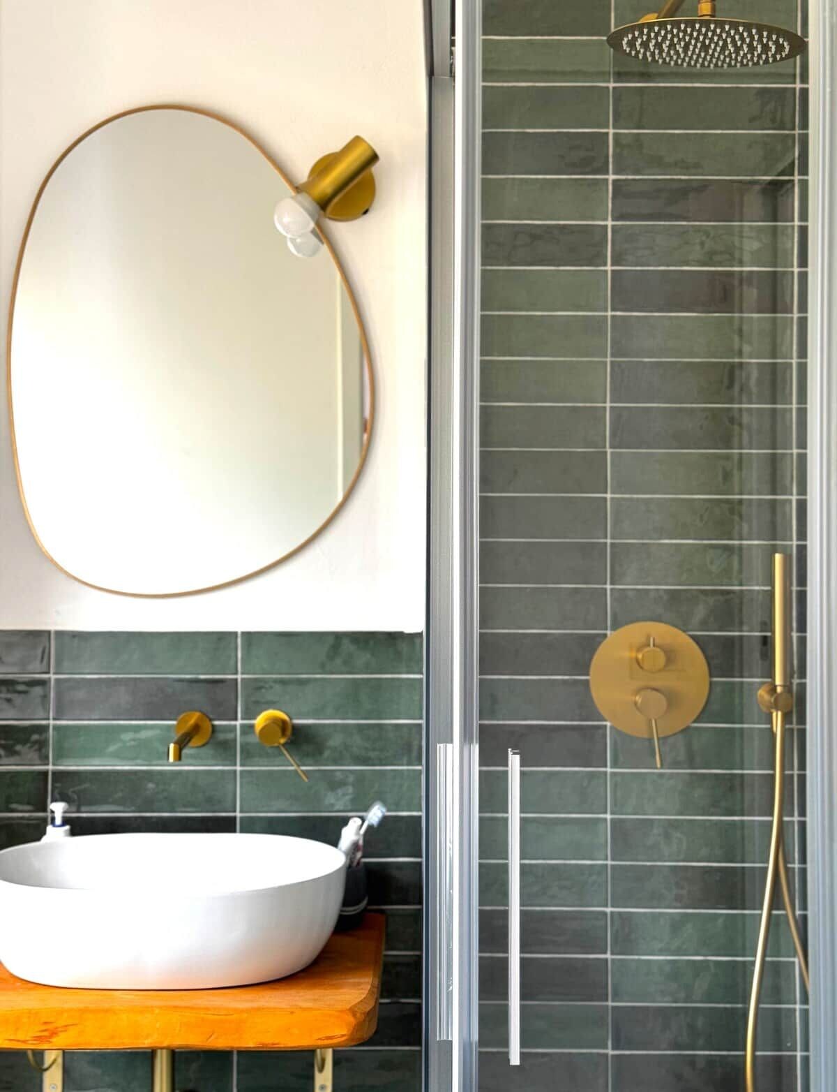 Une salle de bain avec un miroir rond, un lavabo blanc et une douche dotée d’un pommeau de douche doré.
