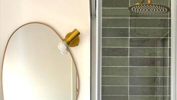 Une salle de bain avec un miroir rond, un lavabo blanc et une douche dotée d’un pommeau de douche doré.