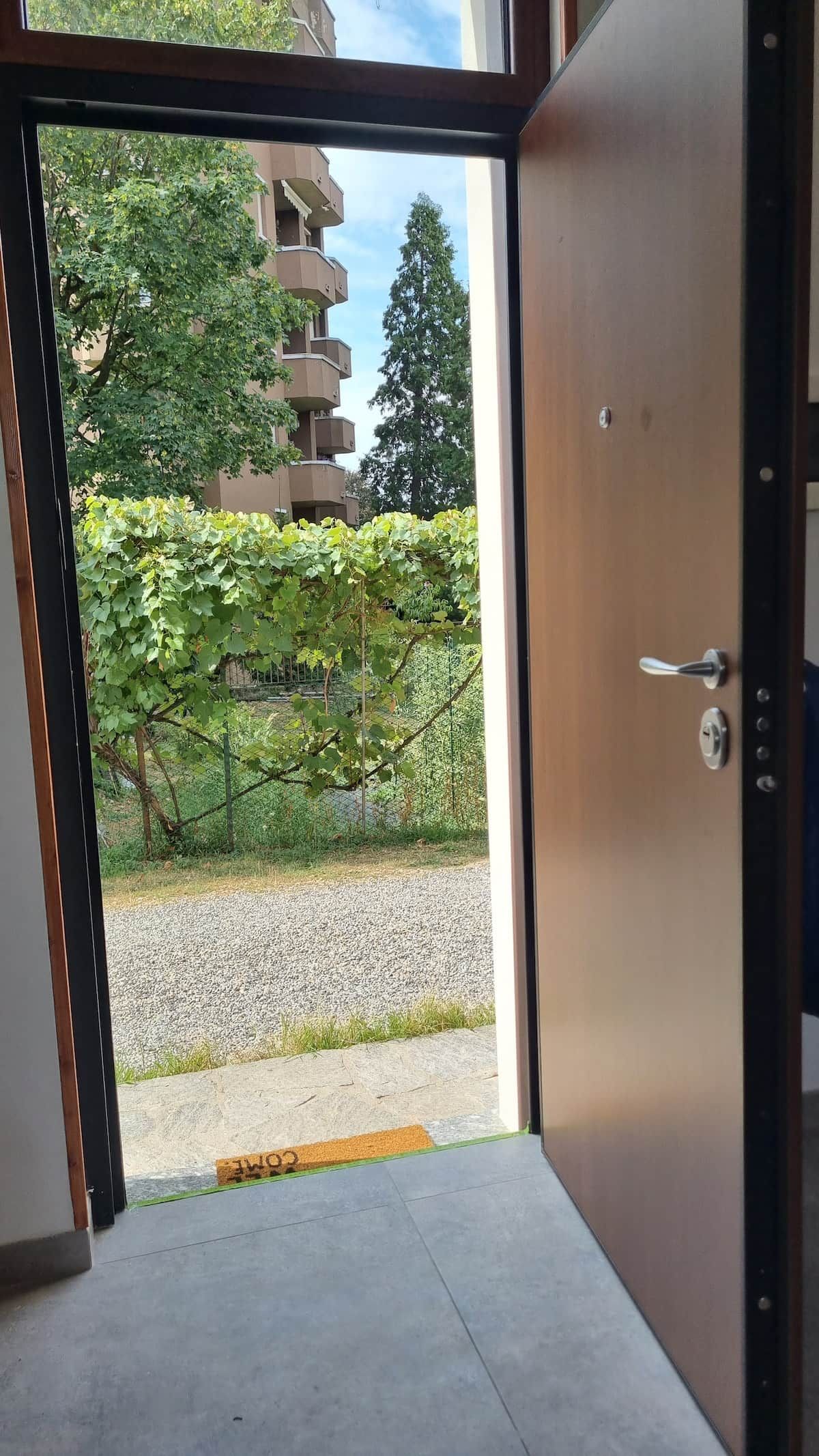 Une porte ouverte donne sur une cour intérieure, où l’on aperçoit un chemin de gravier, une clôture en métal et, au fond, un bâtiment doté de plusieurs balcons.