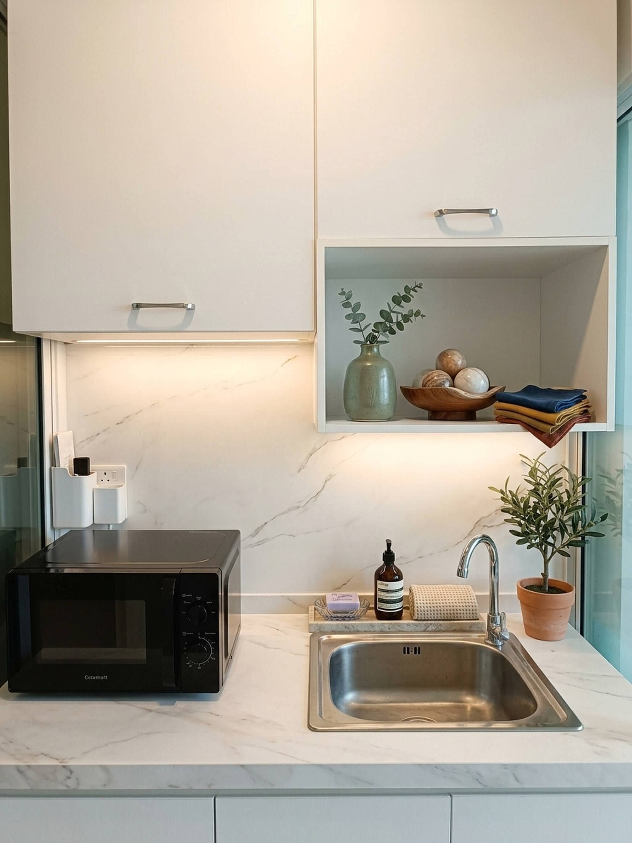 A modern kitchen with a built-in microwave, a stainless steel sink, and a shelf above the sink containing a vase, a bowl of fruits, and folded towels.