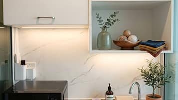 A modern kitchen with a built-in microwave, a stainless steel sink, and a shelf above the sink containing a vase, a bowl of fruits, and folded towels.