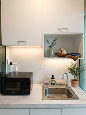 A modern kitchen with a built-in microwave, a stainless steel sink, and a shelf above the sink containing a vase, a bowl of fruits, and folded towels. - Room-regent Home Bang Na Condo- Private (Bangkok)