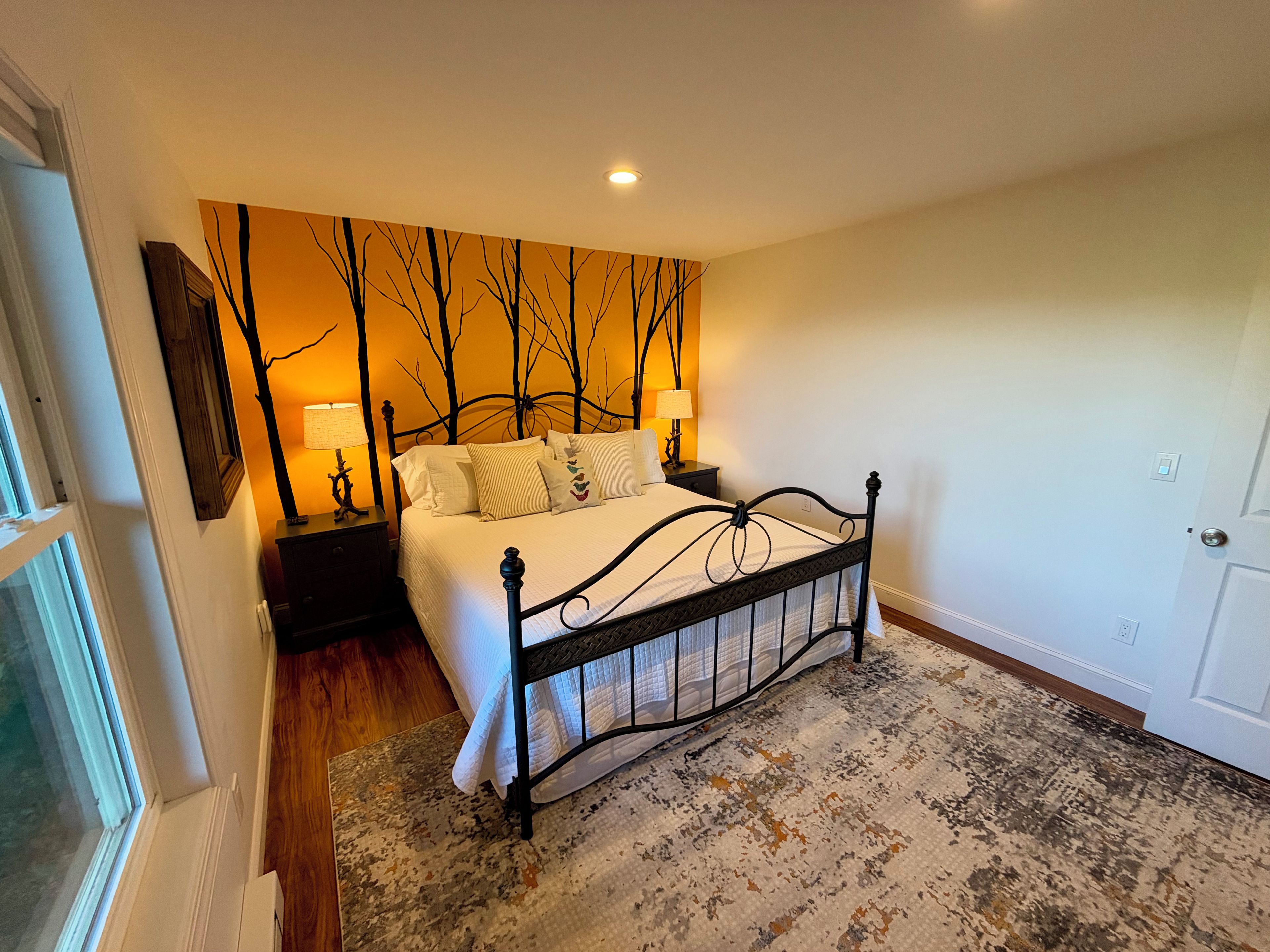A bedroom with a bed, nightstands, a wall mural, and a patterned rug.